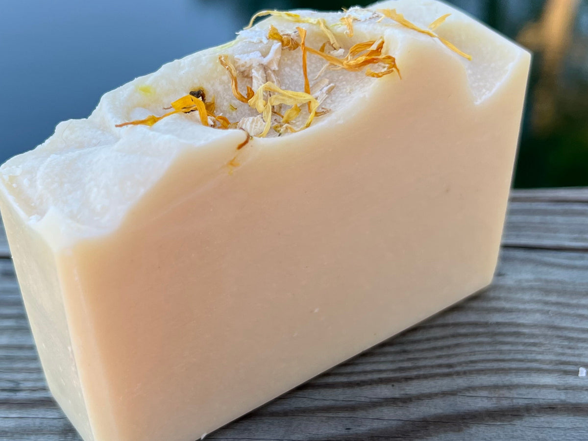 A rectangular bar of pale peach-colored soap with dried orange flower petals on top, resting on a weathered gray wooden surface.